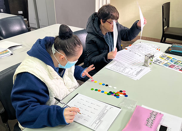 Two people are sitting at a desk, one person is using plastic counters, the other person is holding up some work sheets