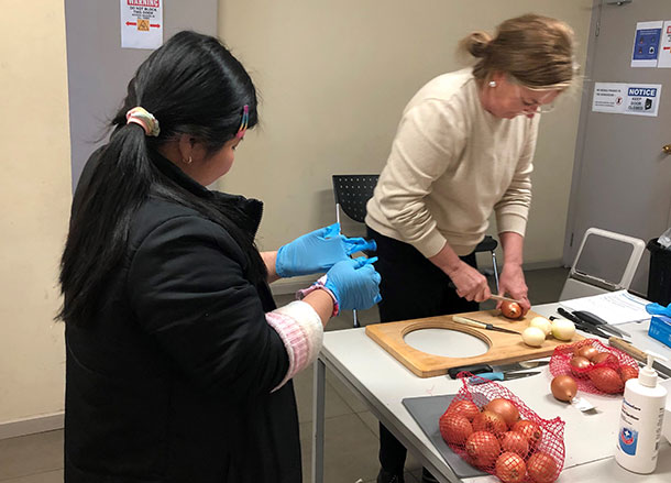 Two people stand at a kitchen table that has net bags of onions on it, one person is chopping onions on a chopping board the other is putting on blue latex gloves