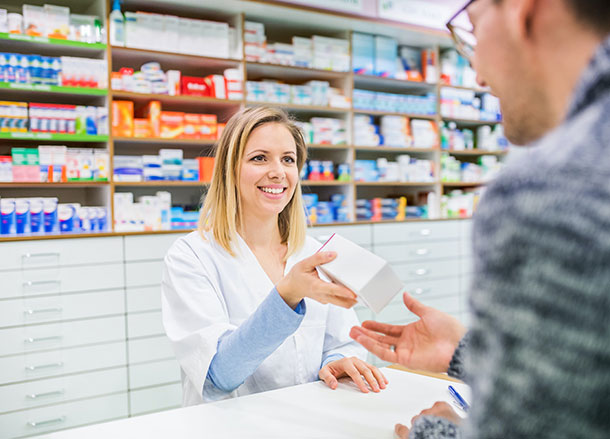 A pharmacist stands in front of shelves of medication, she is passing a box over to a customer