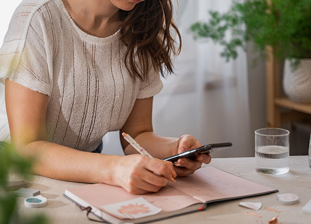 A woman sits at a table holding a phone in one hand and writing in a note book with the other hand