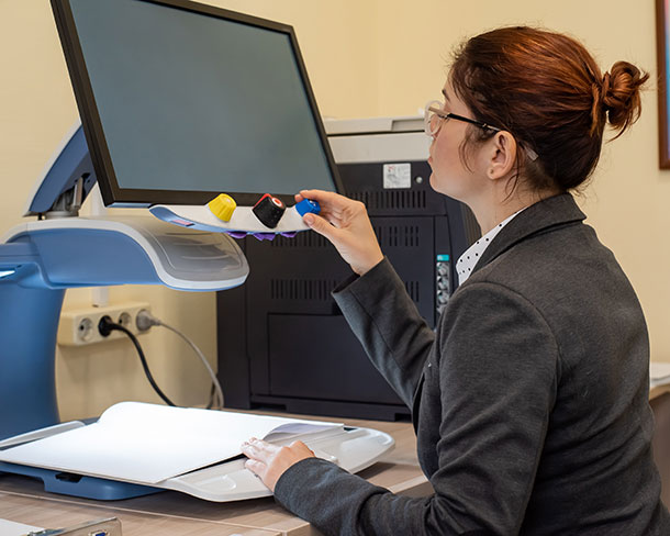 A woman wearing a blazer is seated at a desk using assistive technology to read a document