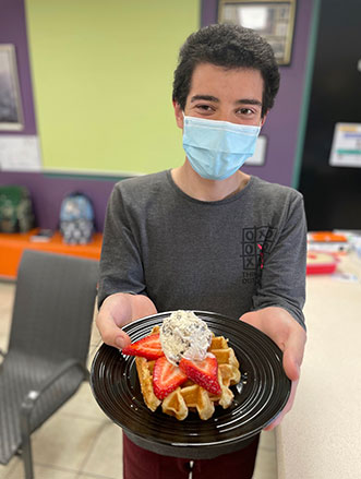 Ruben is wearing a mask, he is holding out a plate of waffles with strawberries and ice cream