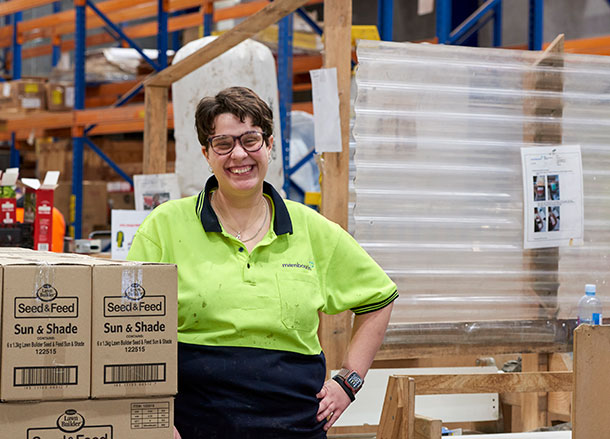 A young woman is standing at a workstation in a warehouse, she is wearing a high-vis work shirt and smiling