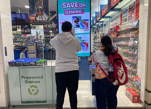 Two people lean against the window of a gaming store, they are holding up pen and paper to take notes