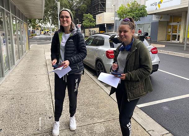 Two young women stand in a retail strip, they are holding notepads and pens in their hands