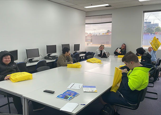 A group of SLES participants sit around a long board table, they are all looking into bright yellow tote bags