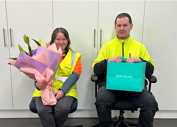 Sarah and Sean are sitting on office chairs, Sarah is holding a large bunch of flowers and Sean is holding a paper gift bag from Koko Black