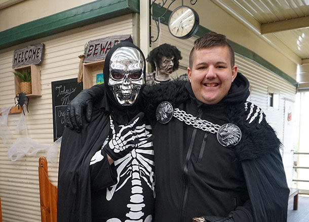 A person dressed in a skeleton costume and mask has their arm around the shoulder of a person with a black cloak, he is smiling