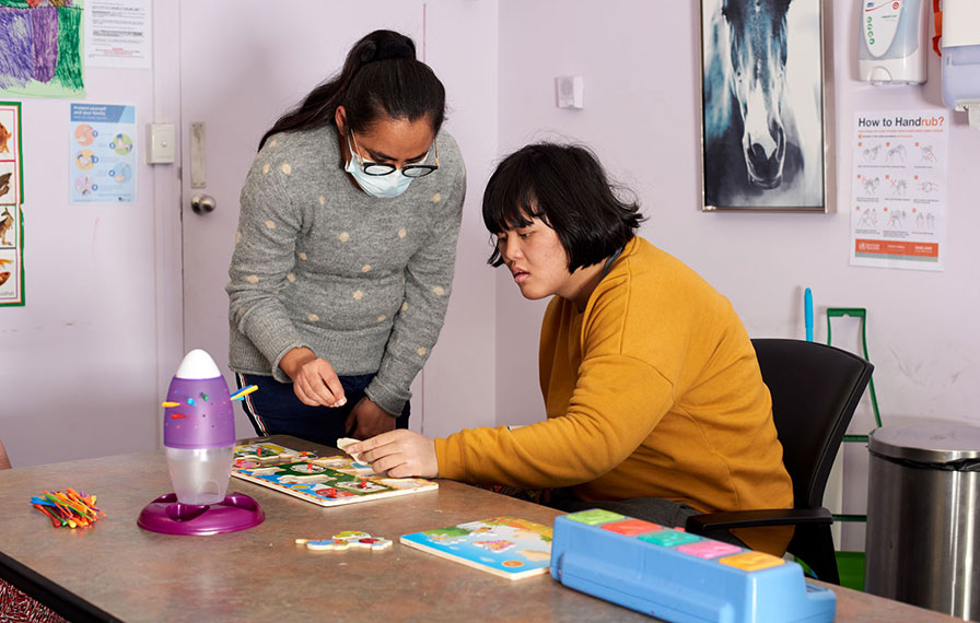 A disability support worker is standing beside a young woman who is seated at a table and working on a puzzle