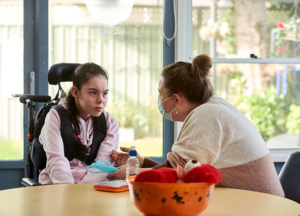 A young woman in sitting in a wheelchair, she is looking at a support worker who is wearing a facemask and leaning in to talk to her