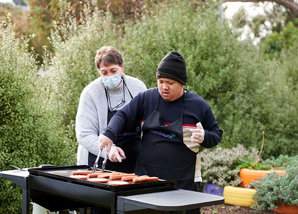 A disability support worker assists a young man to cook sausages on a BBQ