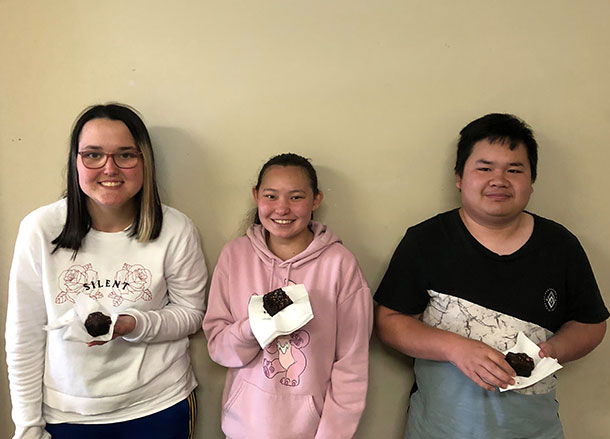 Shakaya, Cindy and Nor are standing against a wall, they are all smiling and holding paper plates with cake on them