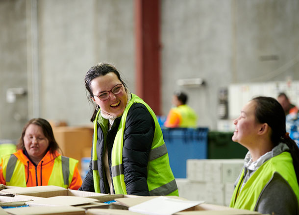 Shakaya is standing at a work station in a warehouse, she is laughing with a colleague standing beside her