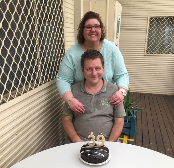 Shane is sitting with Susan standing behind him with her hands on his shoulders, they are both smiling, there is a cake on the table in front of Shane with candles in the shape of 39