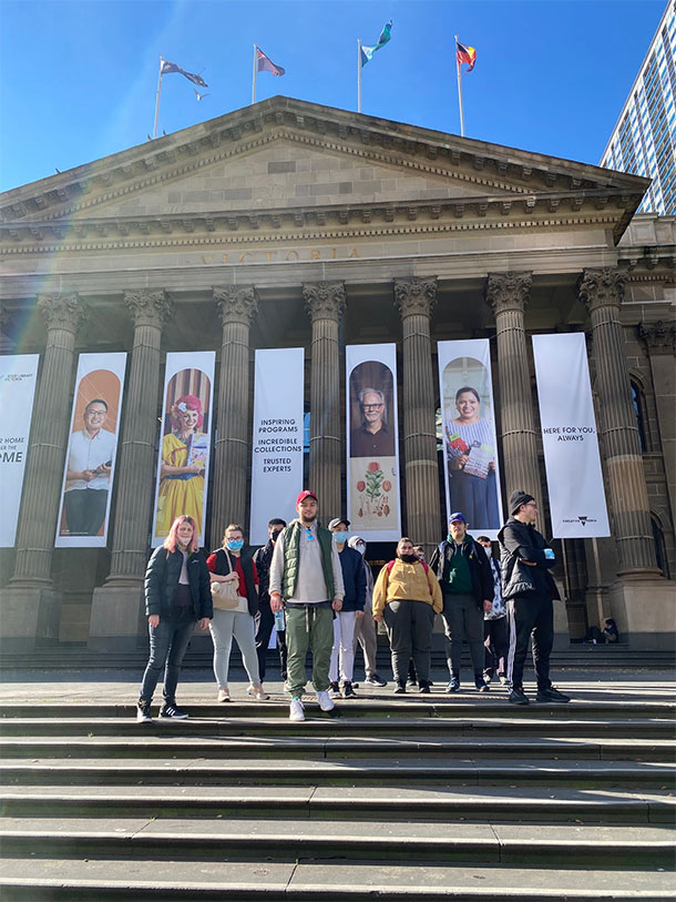 11 people stand on the steps in front of the State Library, there are large banners hanging from the building