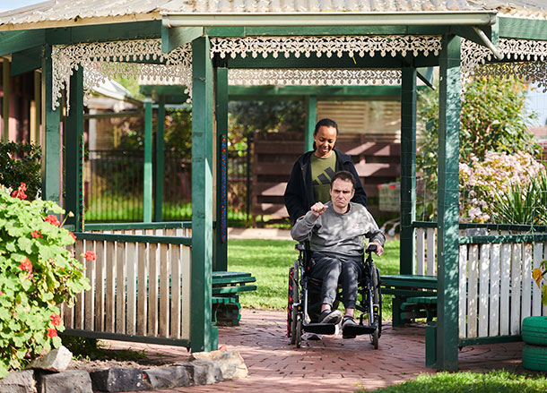 A woman wheels a man in a wheelchair through a garden