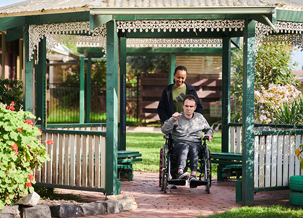 A woman wheels a man in a wheelchair through a garden 