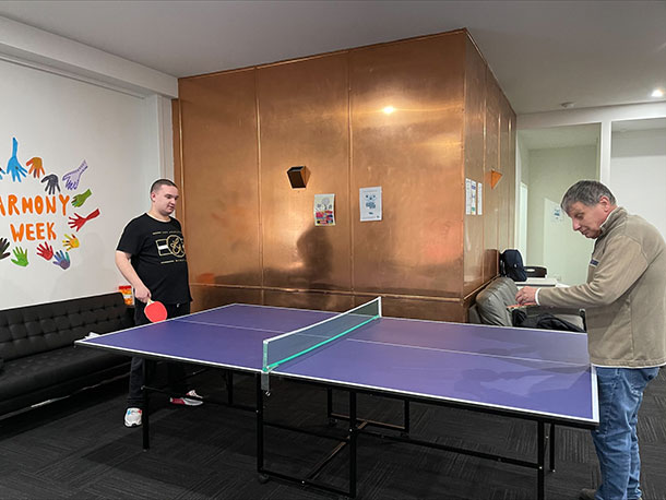 Two people stand at a table tennis table with bats in their hands, one person is holding the ball about to serve