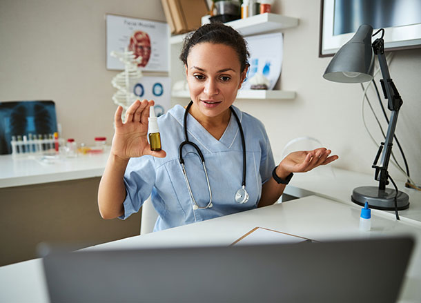 A doctor is sitting in her office holding up some medication to a laptop screen