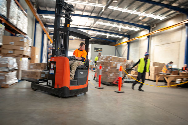 A trainee is moving pallets using a forklift. Another trainee pulls a pallet on a trolley.