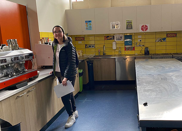 A young woman stands in a hospitality training kitchen in front of a commercial coffee machine