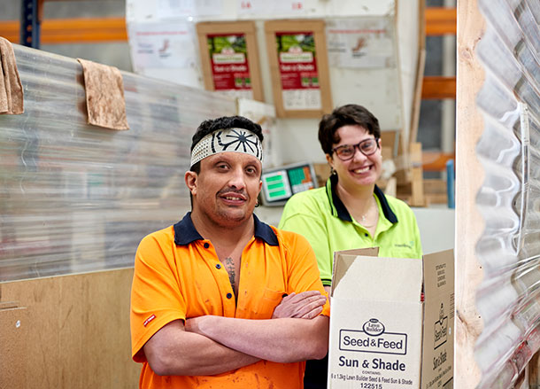 Two people in high-vis workshirts stand at a workstation next to each other, they are both smiling