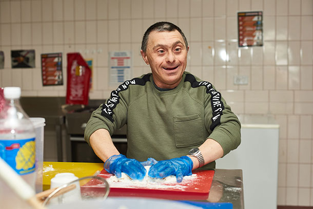 A man wearing latex gloves kneads flour on a chopping board, he is smiling