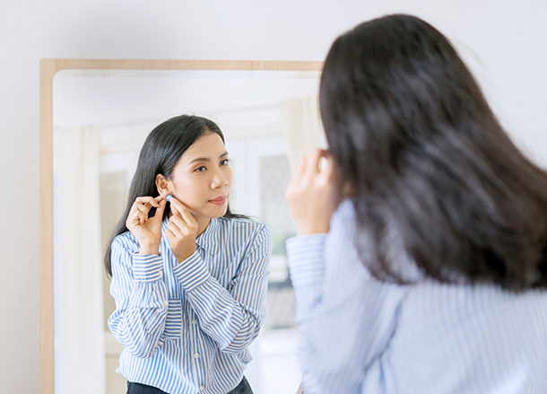 A young woman wearing a business shirt is looking in the mirror and putting on an earring