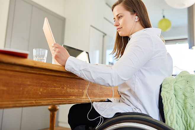 A woman is sitting at a dining table in a wheelchair, there is a laptop on the table, she is holding a tablet and wearing earbuds