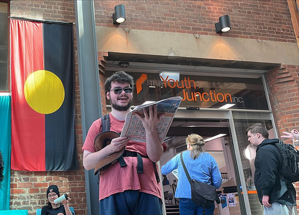 A woman is sitting holding a coffee cup, a man is standing holding a newspaper, two people are about to endter a building with a sign saying Youth Junction