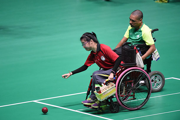 Two people in wheelchairs on a green court marked with white lines, one person has just rolled a boccia ball which is on the court in front of her