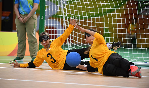 Two people in sports uniform and wearing masks over their eyes are lying in front of a goal net are smiling and raising their arms to stop a ball coming towards them