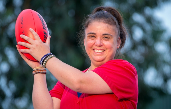 This is an image of a woman holding a footy like she has juts caught it. SHe is smiling at the camera and is wearing a red t-shirt and some beads around her wrist. She is holding a red Australia Rules footbal. There are trees in the backround so she likely standing on an oval. The woman has dark hair pulled back into a ponytail. 