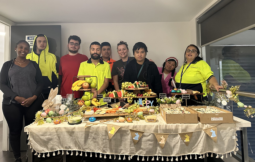 A group of people standing behind a table. The table has a light yellow table cloth with grazing platters on it.