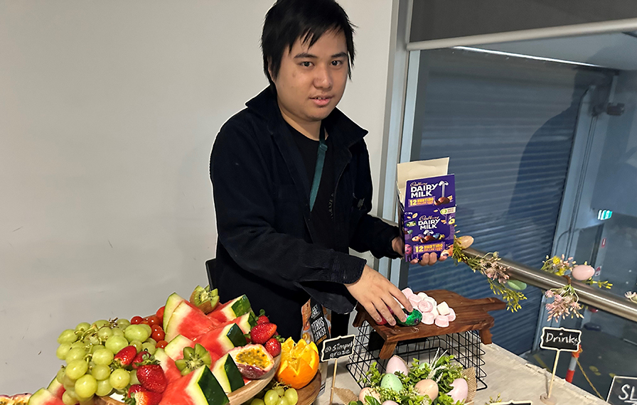 Mikhail stands behind a table and is placing a chocolate Easter egg on a wooden platter. A plate of fruit and Easter decorations are on the table.