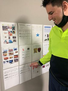 Tristan is standing and pointing at some educational posters on the wall, he is smiling and wearing a high-vis shirt