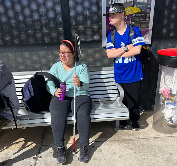 One person is sitting at a bus stop holding a white stick, a second person stands beside her looking out for a bus to arrive