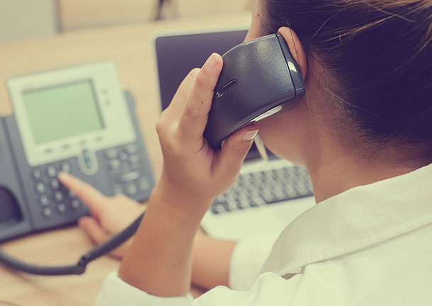 A woman holds a phone receiver to her ear and dials a number on a landline phone on her desk